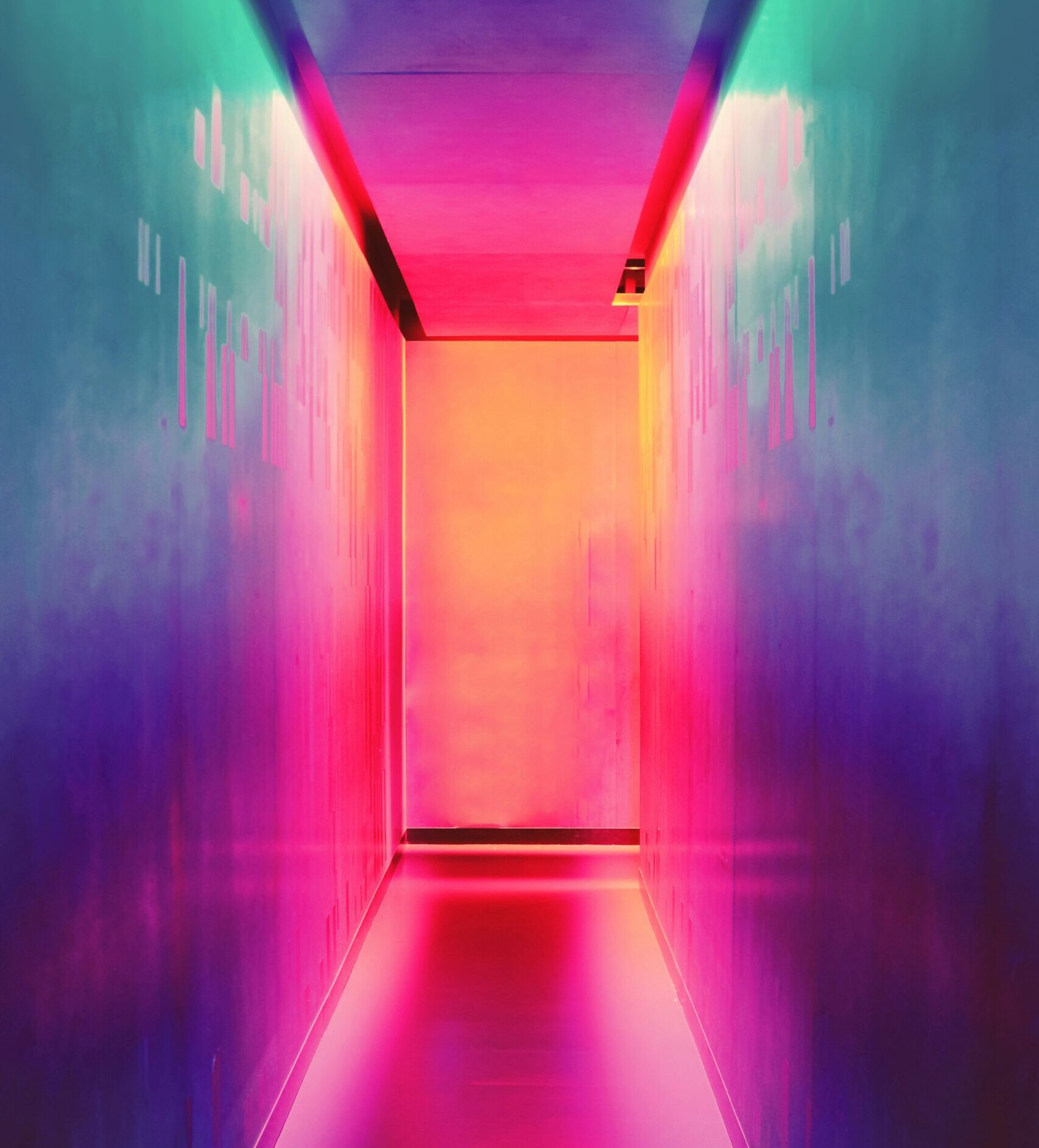 A brightly lit corridor with walls and ceiling illuminated in a gradient of vibrant colors, including shades of pink, orange, purple, and teal. The reflective floor enhances the neon glow, creating a visually striking and futuristic ambiance.